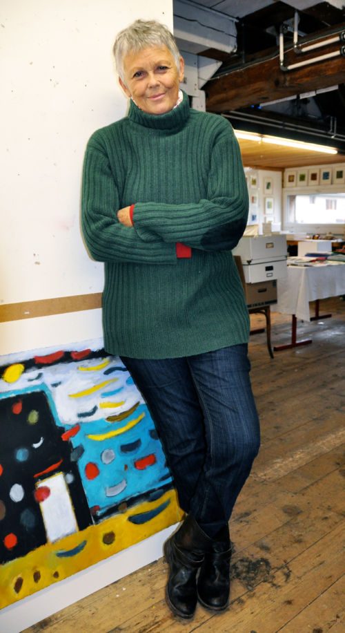 Katharina Rapp mit grünem Rollkragenpullover und verschränkten Armen, an die Wand ihres Ateliers gelehnt, davor ein farbiges Bild
