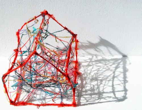 Hausförmiges Objekt aus rot umwickeltem Gerüst mit wirr verspannten bunten Fäden, das einen Schatten an die Wand wirft
