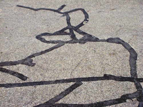 Schwarze, geschwungene Teerlinien auf grauem Asphalt, die wie eine Zeichnung am Boden wirken
