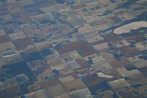 Luftaufnahme einer flachen Landschaft mit rechteckigen Feldern in Braun- und Grautönen und kleinen Seen