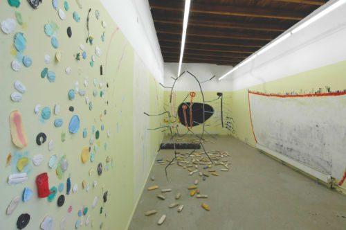 Ausstellungsraum mit farbigen Fundstücken an der Wand, einer grossen Drahtfigur und am Boden verstreuten Objekten