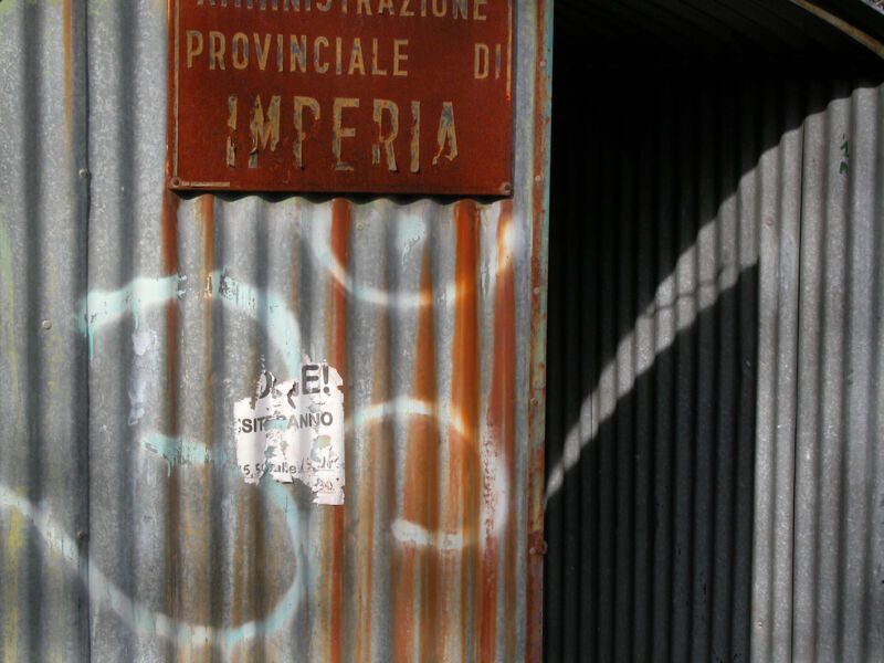 Rostiges Wellblech mit verblasstem Schild «Amministrazione Provinciale di Imperia», weissen Graffitischwüngen und Plakatresten, Fotografie aus der Serie «Alltägliche Fundstücke I»