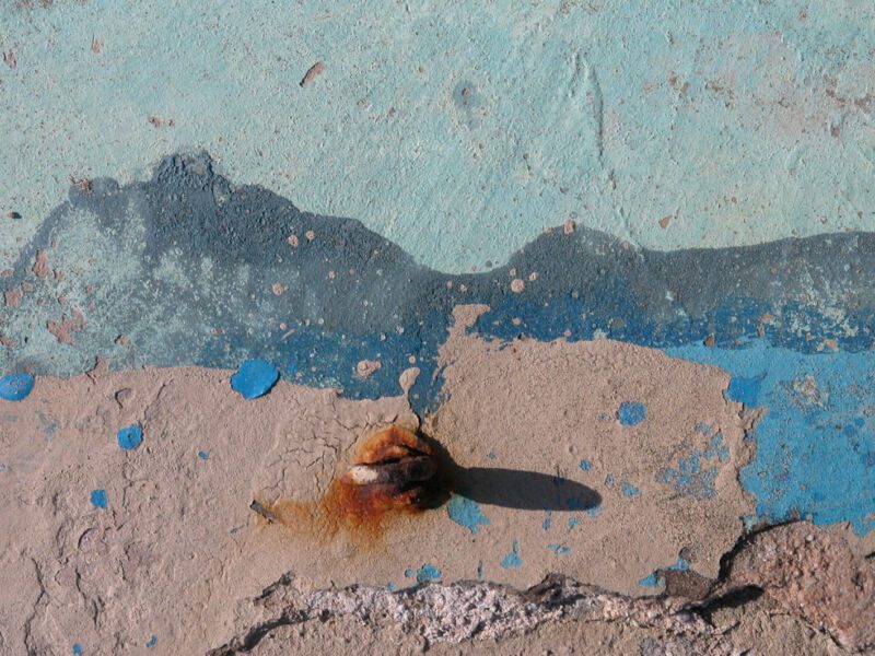 Abgeplatzte Farbschichten in Hellblau, Dunkelblau und Beige mit einem rostigen Metallstummel, der Schatten wirft, Fotografie aus der Serie «Alltägliche Fundstücke I»
