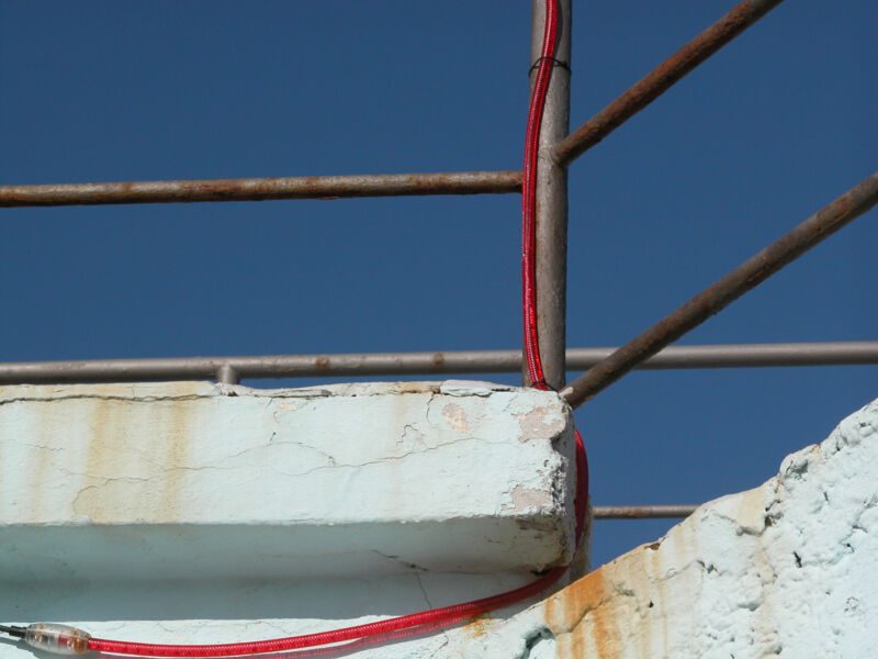 Rostiges Geländer und rotes Kabel an einer weissen, rissigen Betonkante vor tiefblauem Himmel, Fotografie aus der Serie «Alltägliche Fundstücke I»