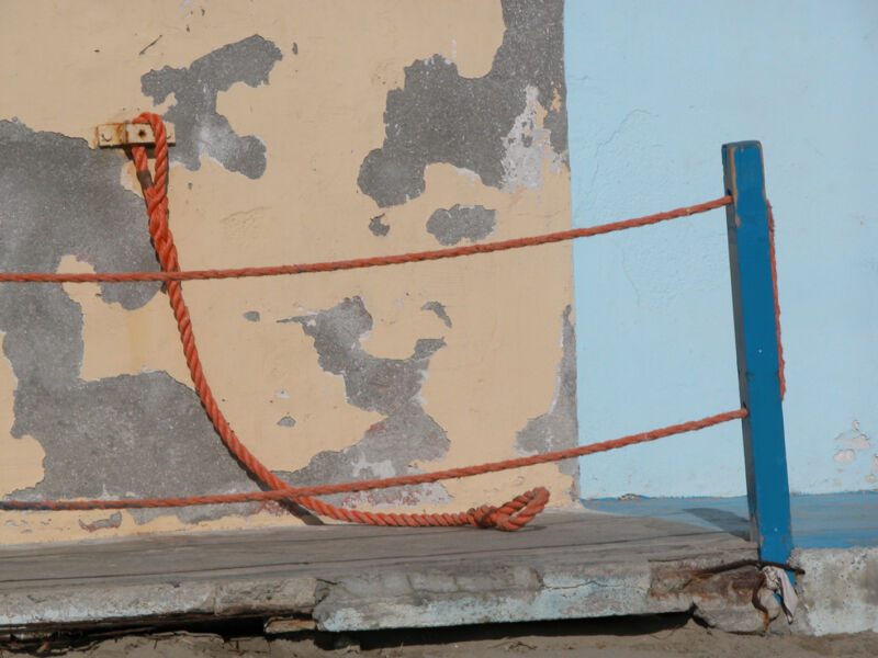 Orangefarbenes Seil zwischen fleckiger Mauer und blauem Pfosten, lose auf einen Holzsteg herabhängend, Fotografie aus der Serie «Alltägliche Fundstücke I»