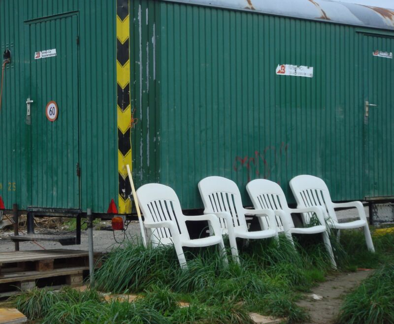 Vier weisse Plastikstühle in einer Reihe im Gras vor einem grünen Baucontainer, Fotografie aus der Serie «Alltägliche Fundstücke II»