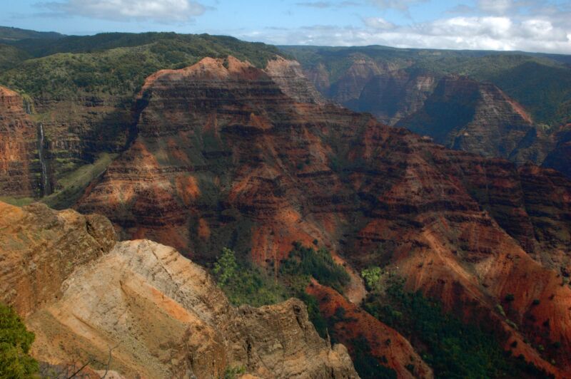 Tief zerfurchte Schlucht mit rot-braunen, grün bewachsenen Felswänden unter hellem Himmel, Fotografie aus der Serie «Hawaii»