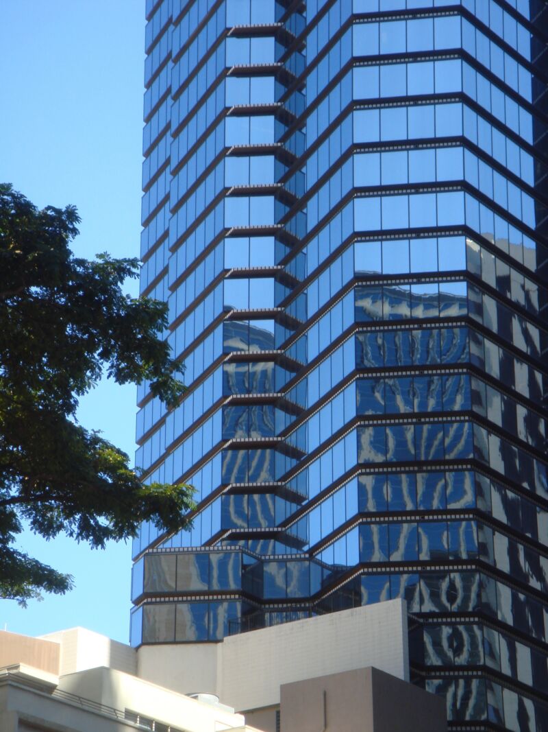 Gestaffelte Glasfassade eines Turms mit wellenförmigen Spiegelungen, links eine dunkle Baumkrone, Fotografie aus der Serie «Honolulu»