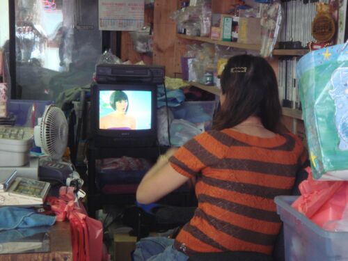 Frau in orange gestreiftem Pullover von hinten vor einem kleinen Fernseher in einem vollgestellten Laden, Fotografie aus der Serie «Honolulu»